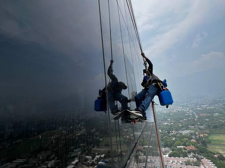 Dos técnicos suspendidos limpiando cristales de una torre con vista a Monterrey